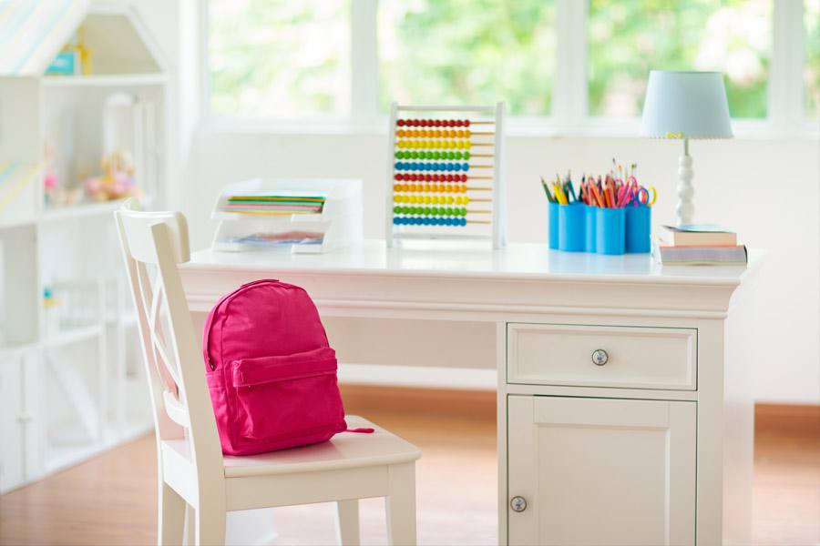 A white student desk and chair with a red backpack on the chair.